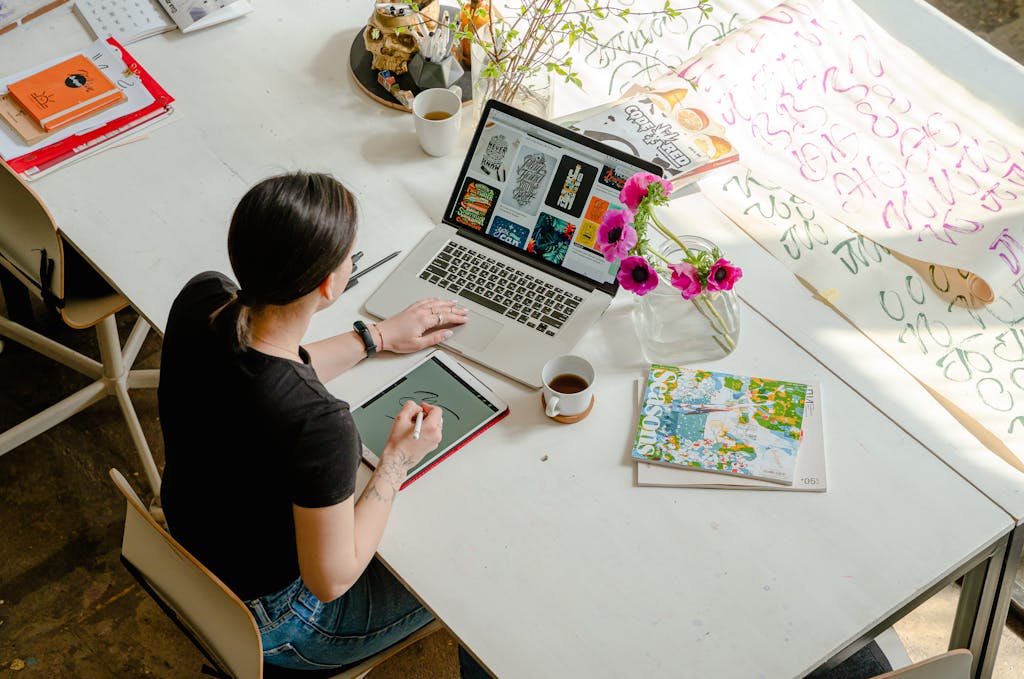 Woman creating digital artwork on a laptop in a sunlit workspace with art supplies.